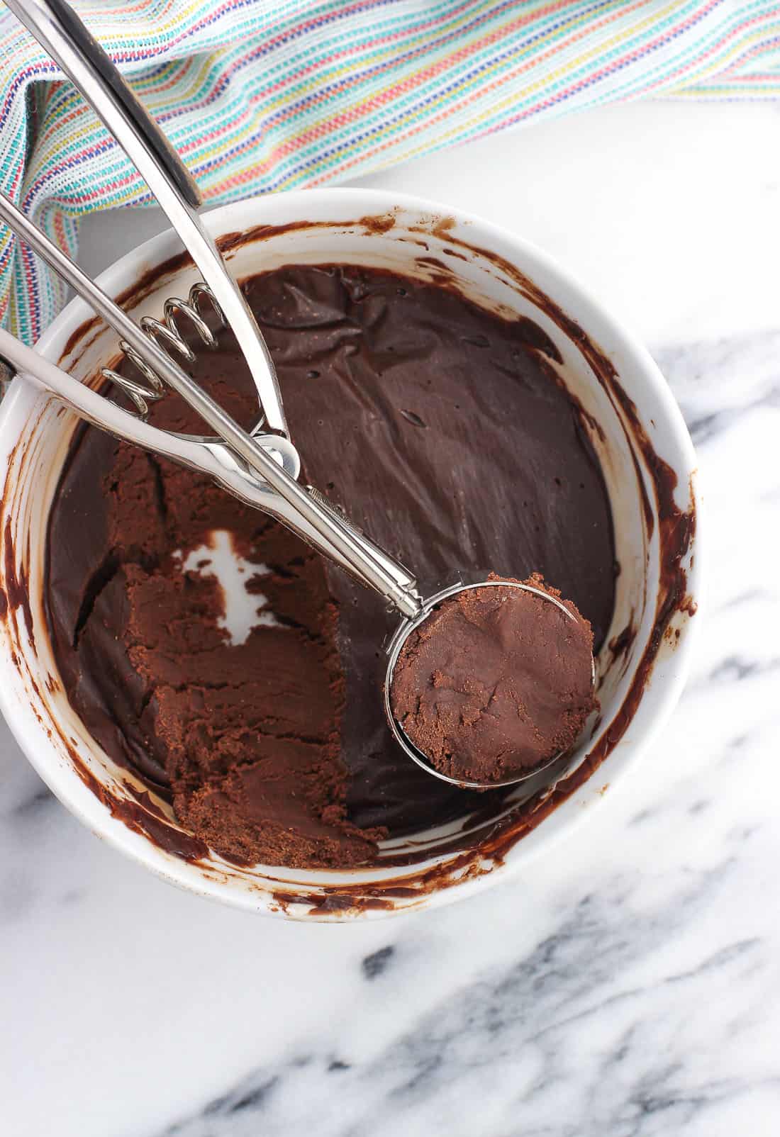 The truffle mixture solidified after being refrigerated in a bowl, with a cookie scoop resting on top after scooping an amount for one truffle