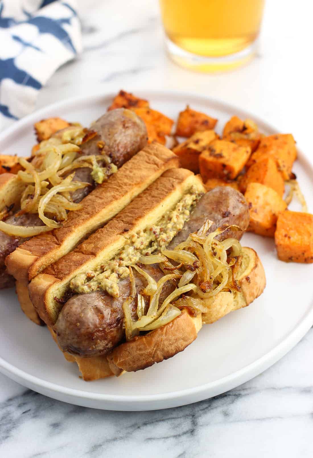 Two bratwursts in buns with the fixing on a plate with roasted sweet potatoes.