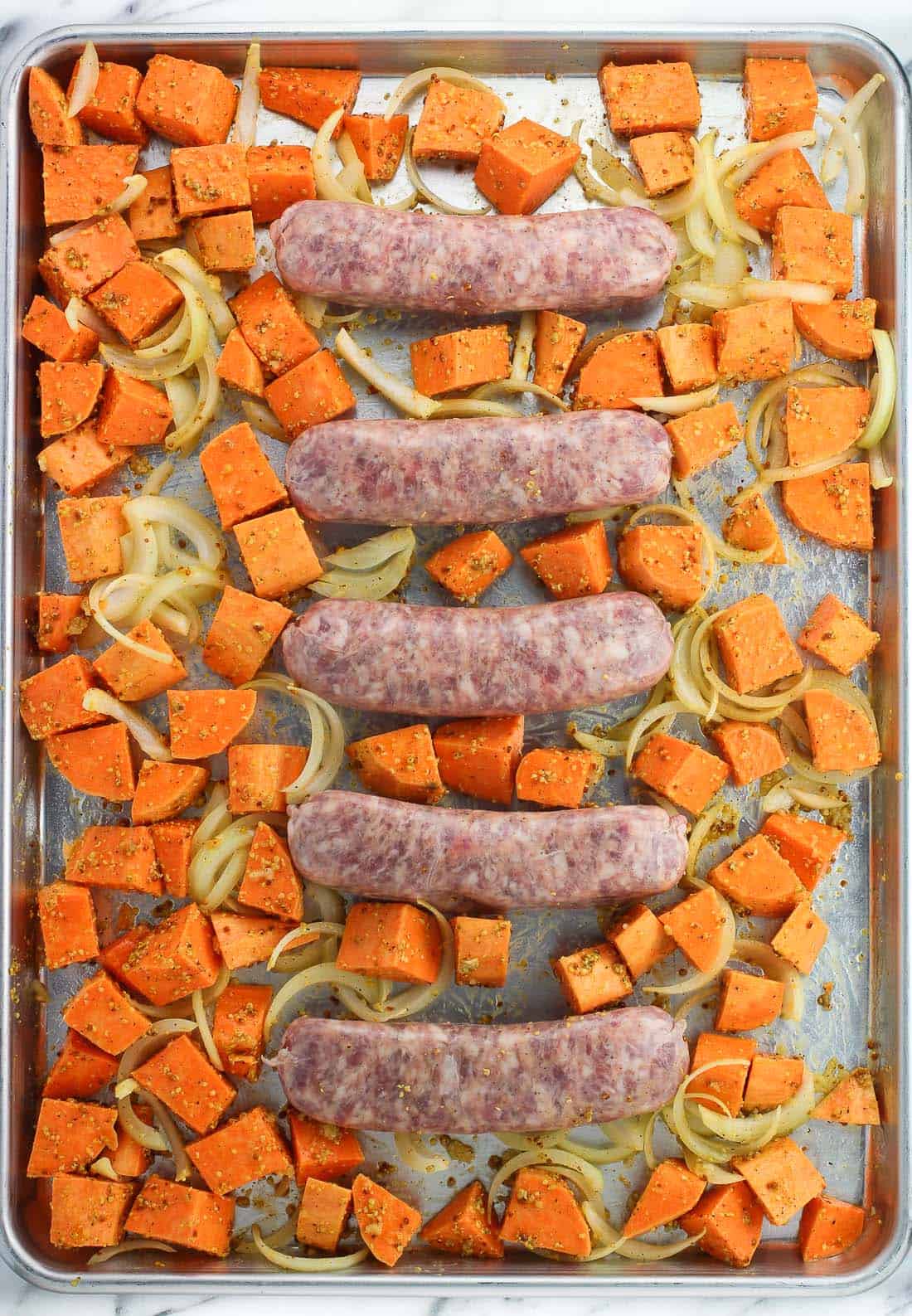Raw bratwursts, sliced onion, and sweet potato chunks on a rimmed sheet pan.