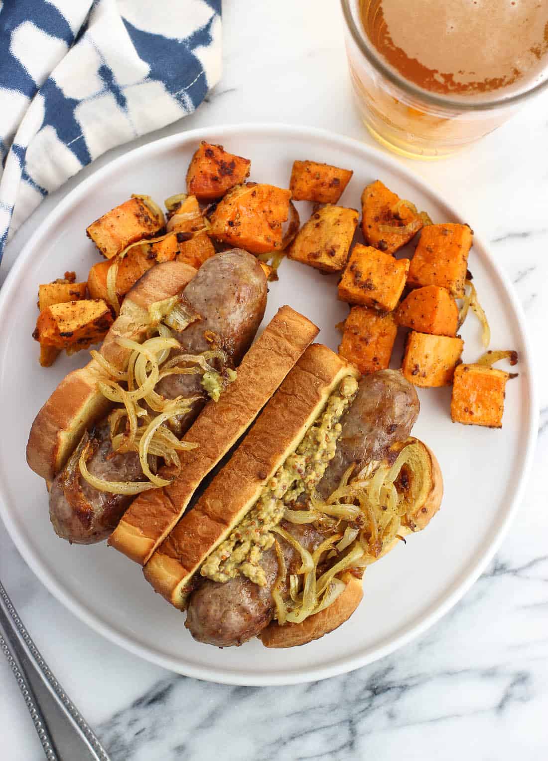 Two bratwursts and sweet potatoes on a plate next to a glass of beer.