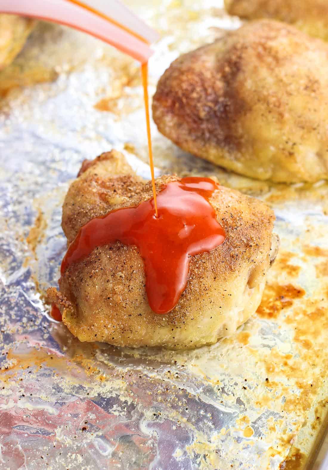 A thin stream of buffalo sauce being drizzled onto a cooked chicken thigh.
