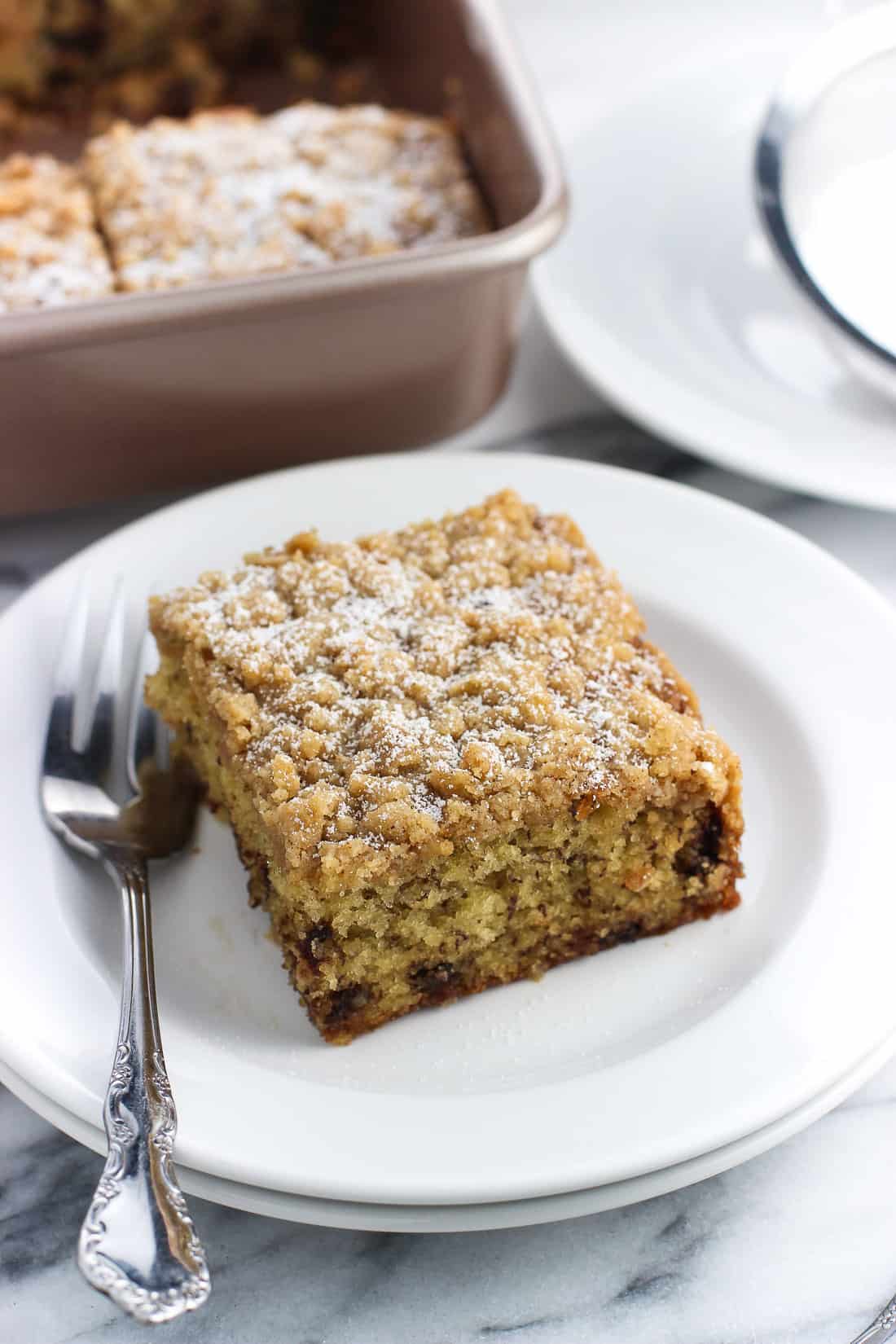 A piece of crumb cake dusted with confectioners' sugar on a small dessert plate next to a fork