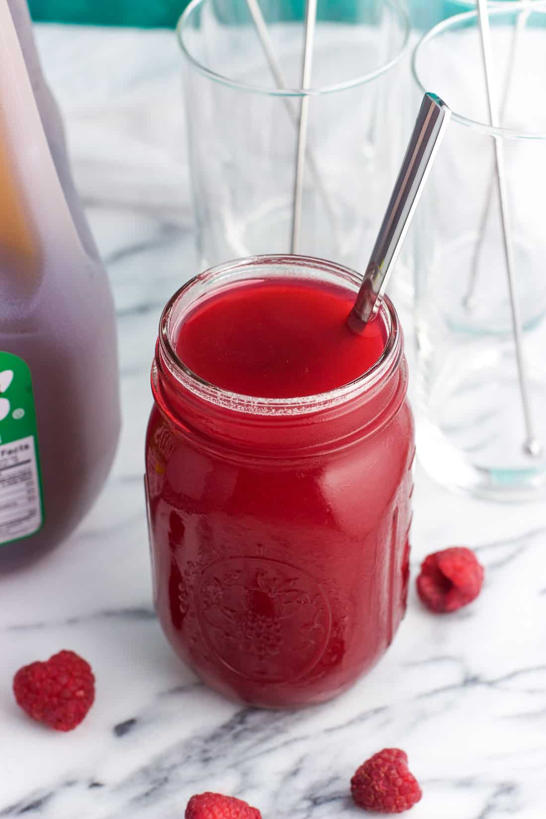 A jar of raspberry simple syrup with a spoon in it next to a gallon of iced tea and tall glasses for serving
