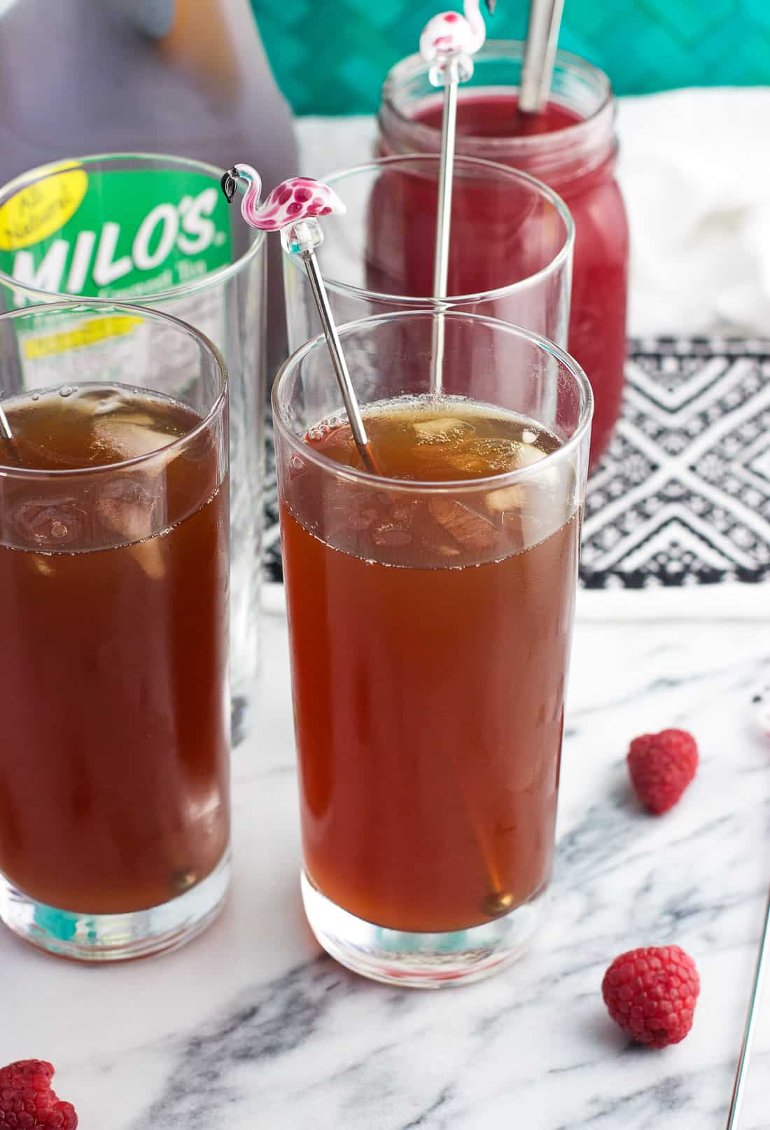 Tall glasses of iced tea garnished with fresh raspberries and flamingo drink stirrers with a jar of raspberry syrup in the background.