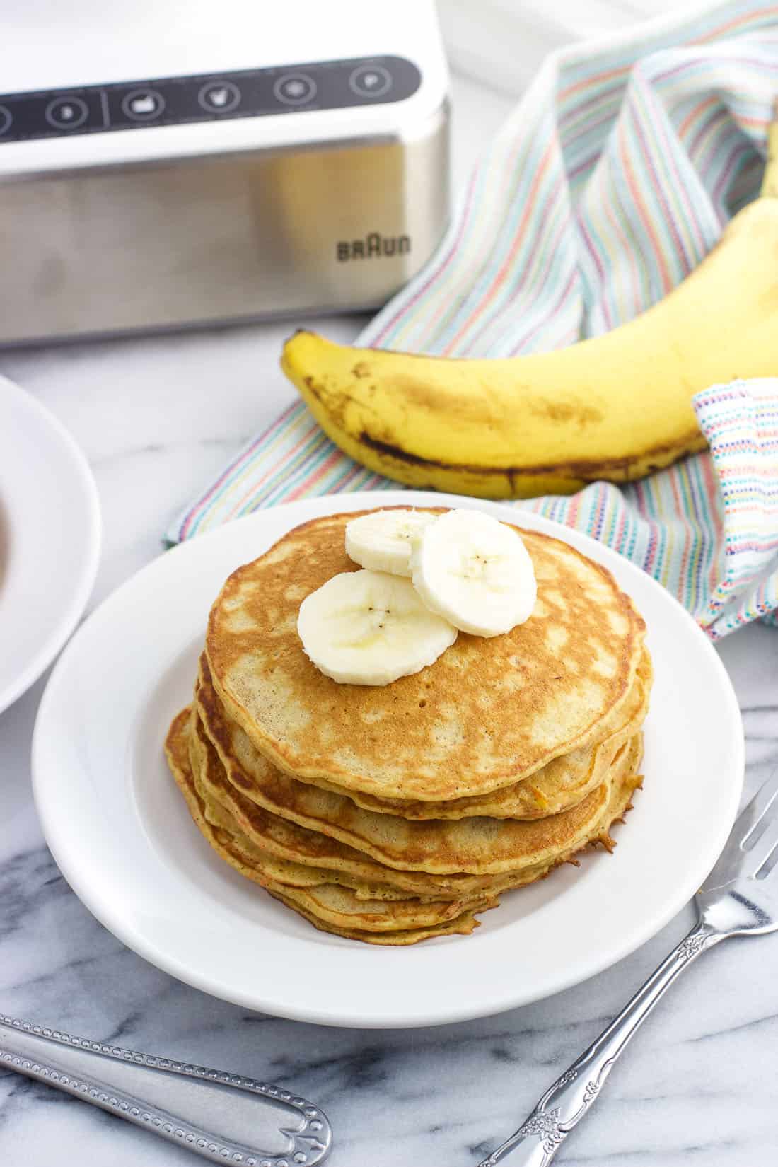 A stack of pancakes on a dish topped with banana slices with a whole banana and a blender base in the background 