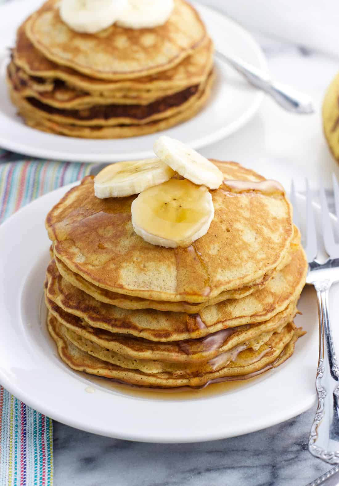 A stack of pancakes topped with maple syrup and banana slices on a small plate, with another plate of pancakes in the background