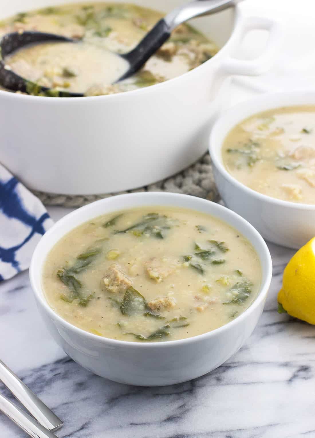 Two dinner bowls of Italian wedding soup in front of a large stoneware pot of soup with a ladle in it