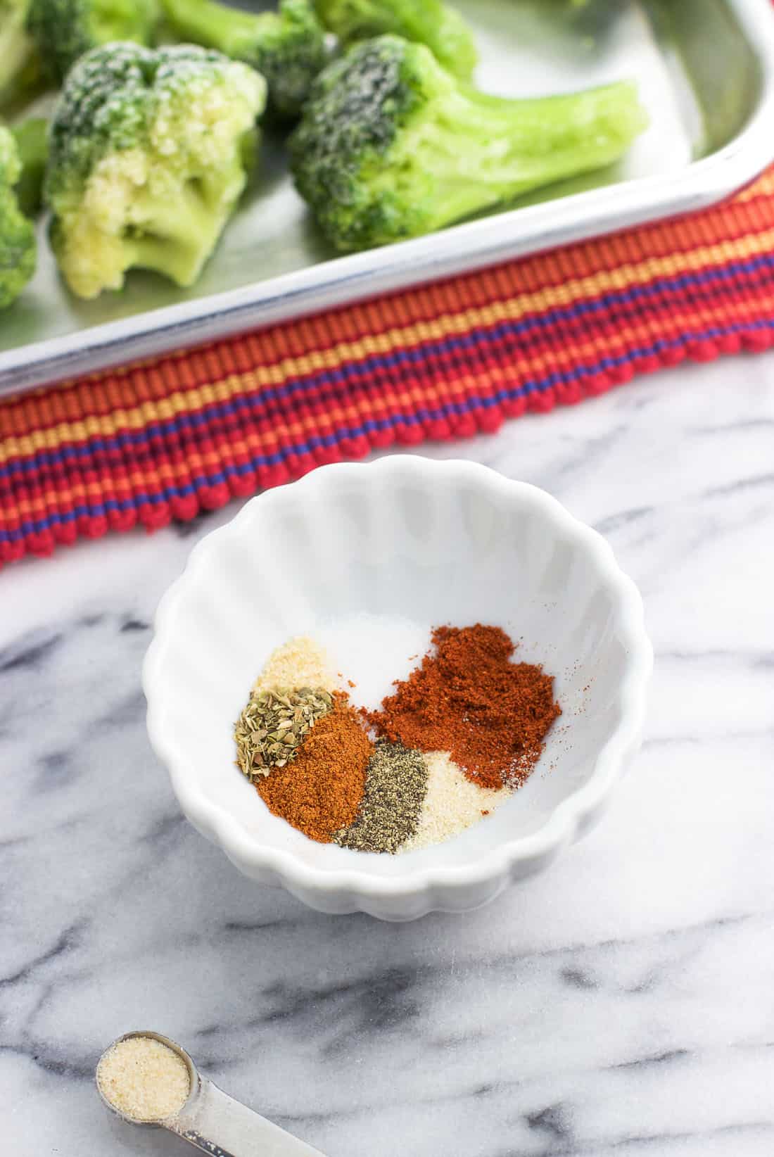 Seasoning mix in a small bowl with frosty broccoli florets on the baking sheet in the background