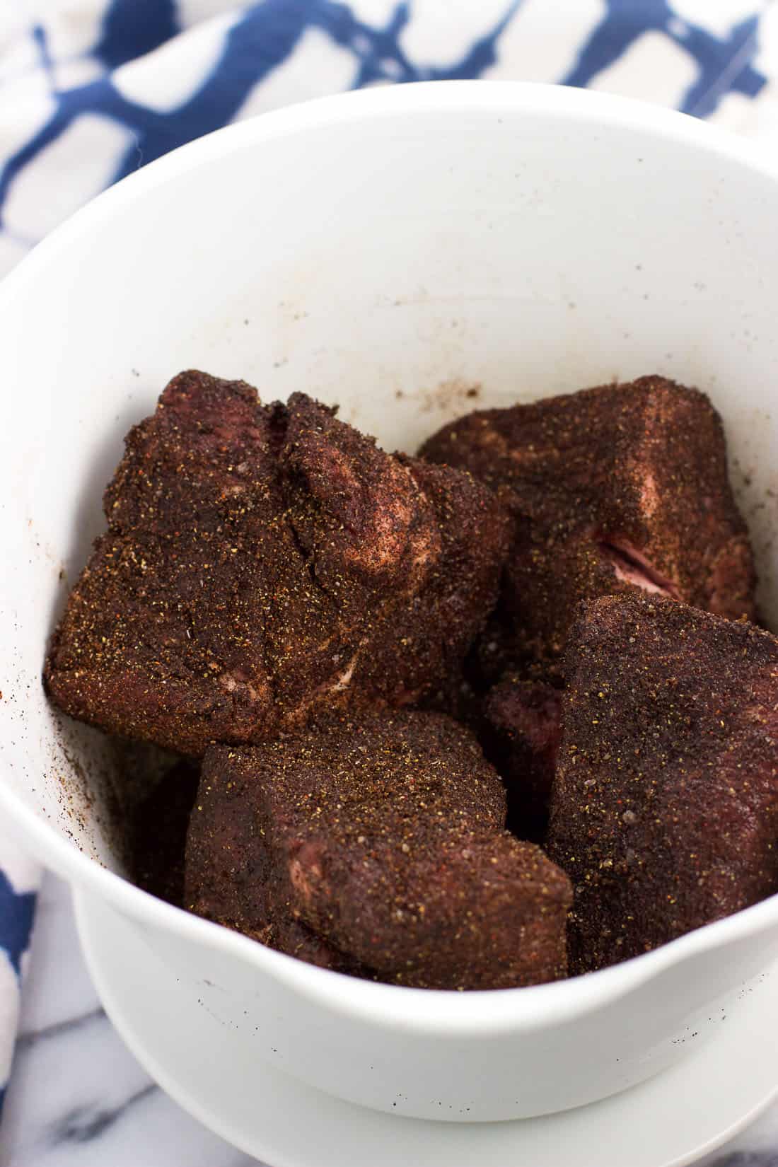 Large chunks of pork roast rubbed in a coffee and spice mixture in a medium plastic mixing bowl.