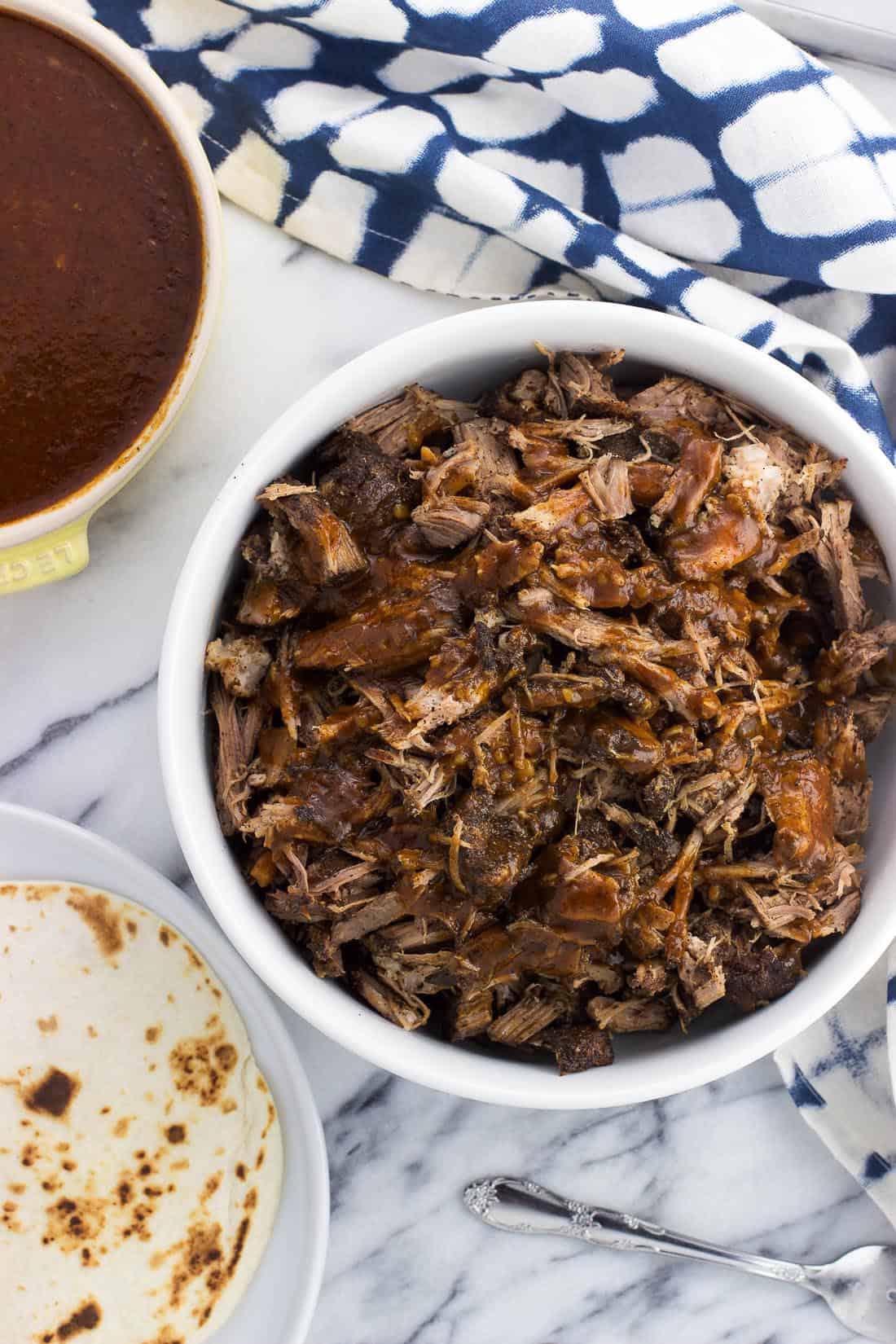 Pulled pork in a serving bowl next to a bowl of homemade BBQ sauce and warmed tortillas.