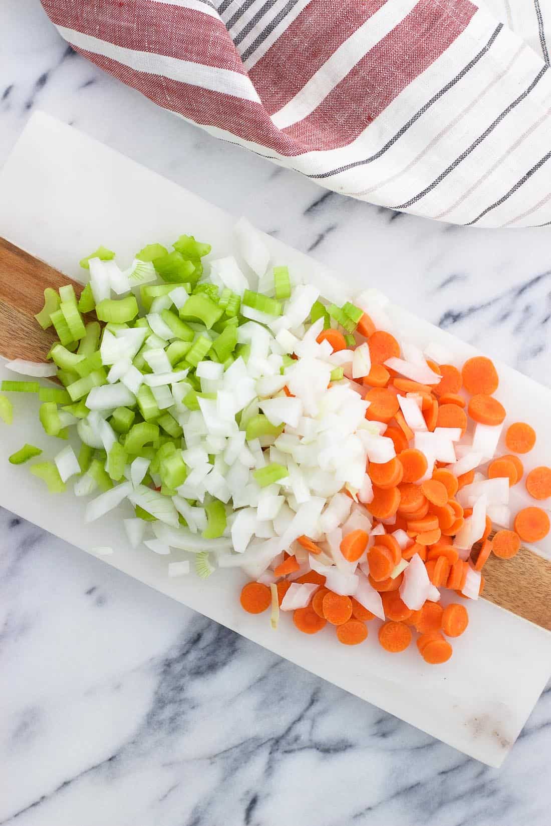 Apio, cebolla y zanahorias picados en una tabla junto a un paño de cocina