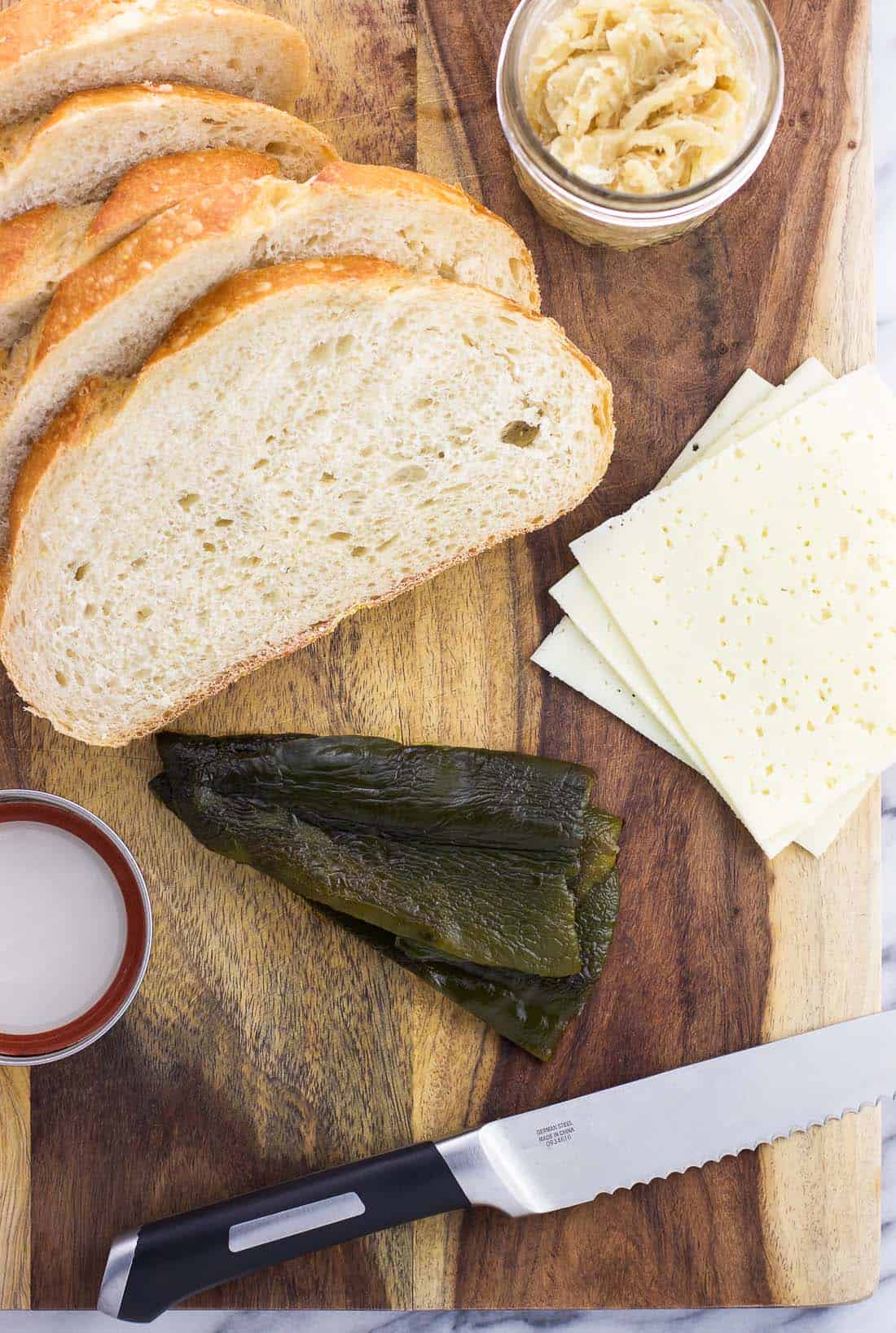 Slices of bread, slices of cheese, roasted poblano pepper strips, and a jar of caramelized onions on a wooden cutting board
