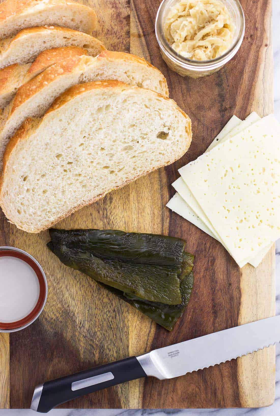 Slices of bread, slices of cheese, roasted poblano pepper strips, and a jar of caramelized onions on a wooden cutting board.