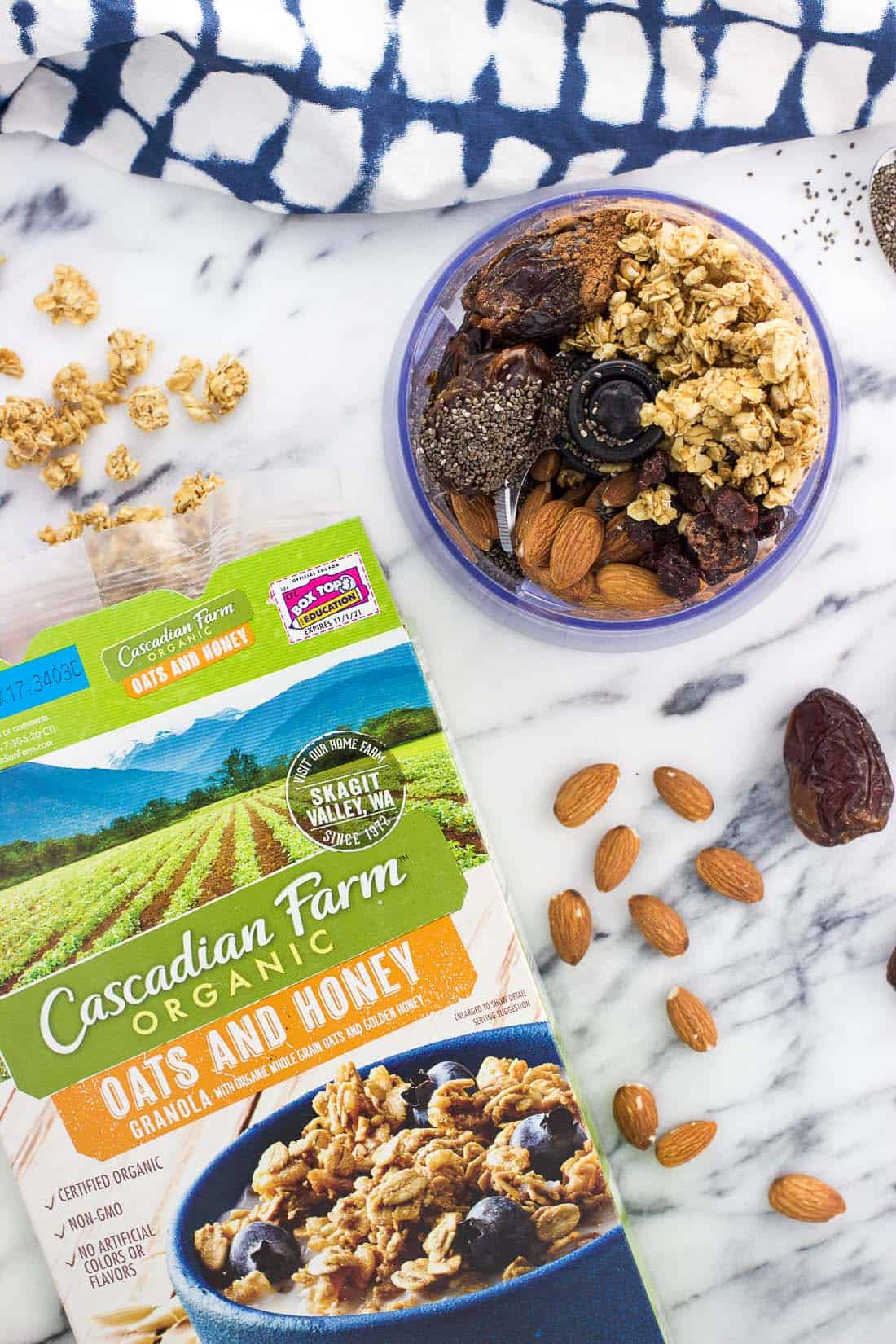 An overhead picture of the recipe ingredients in a small food processor and an open box of granola with granola clusters spilling out onto the board