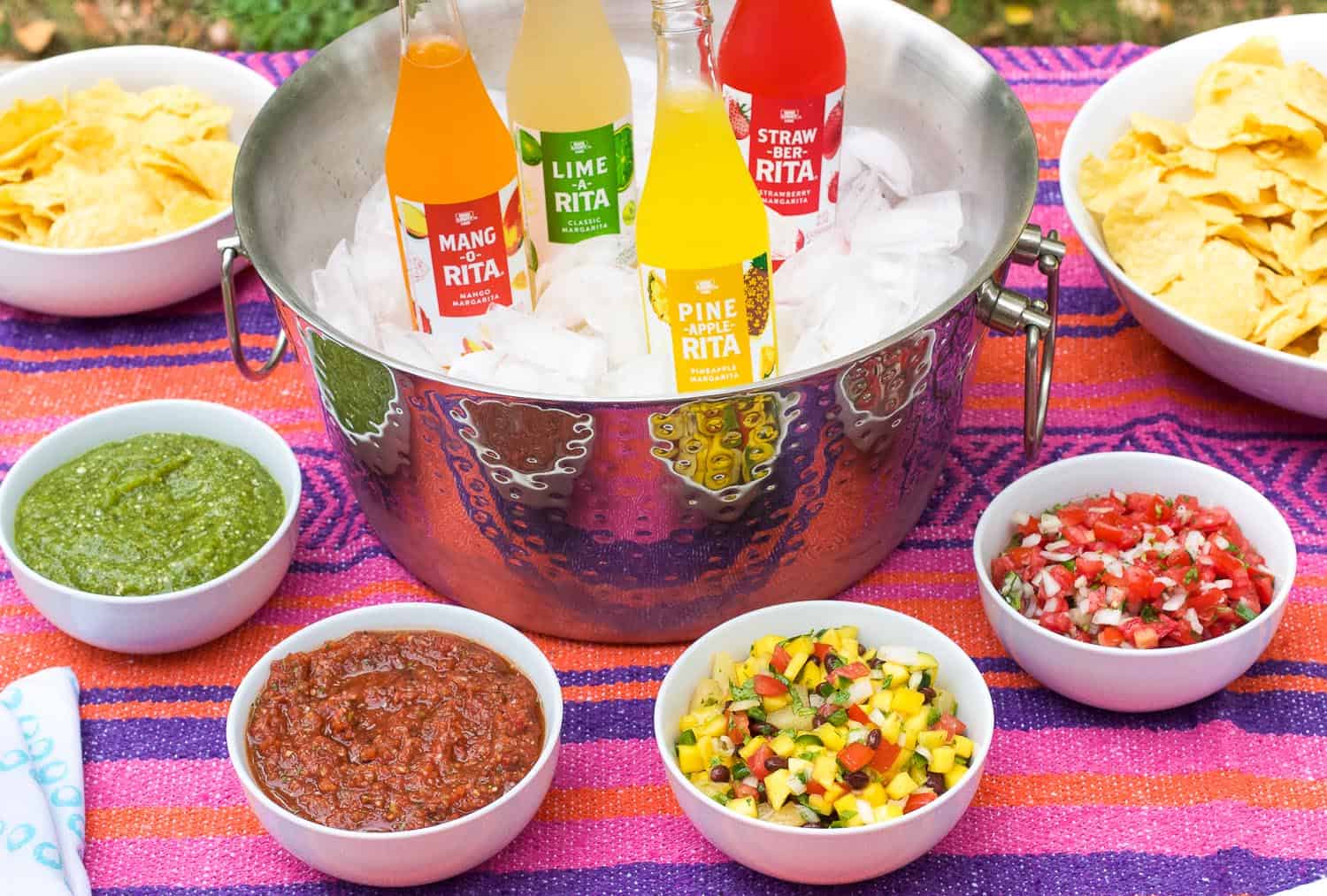 An outdoor table spread of four different salsa, two bowls of tortilla chips, and an ice cooler stocked with RITAS