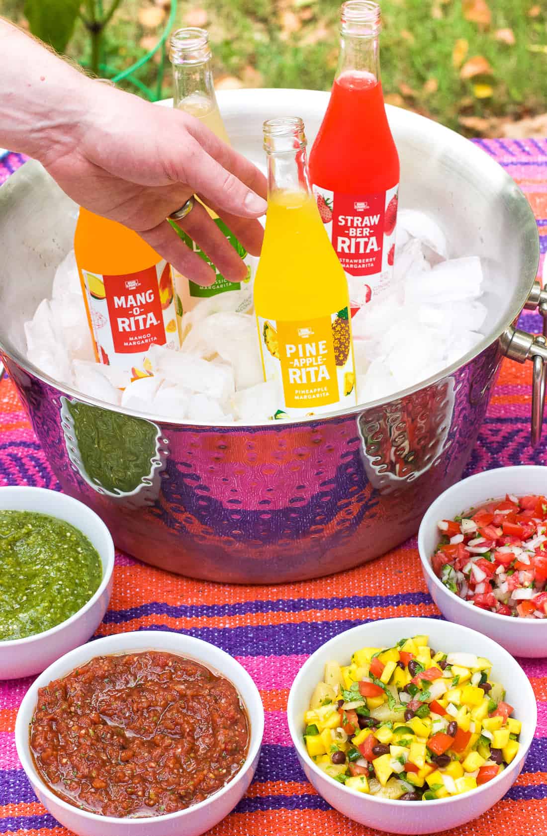 A hand reaching for a RITA in an ice-filled chest surrounded by bowls of tortilla chips and salsas