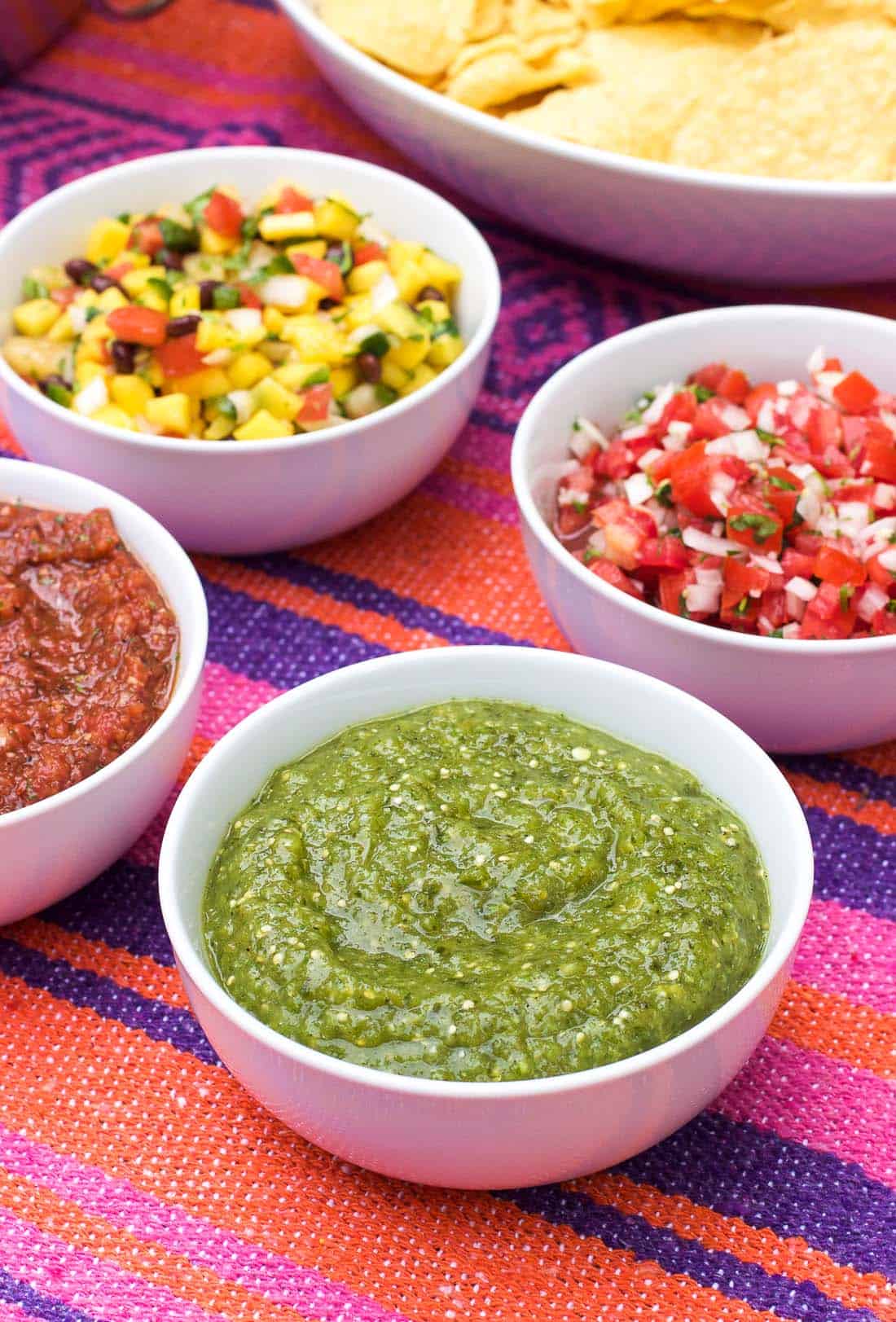 Four bowls of different salsas on a table next to a large ceramic serving bowl of tortilla chips