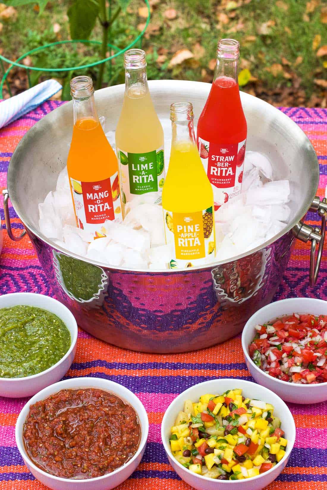 An outdoor table spread of four different salsa, two bowls of tortilla chips, and an ice cooler stocked with RITAS