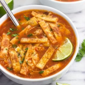 A ceramic bowl of soup topped with tortilla strips, cheese, and lime wedges