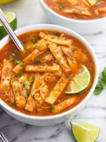 A ceramic bowl of soup topped with tortilla strips, cheese, and lime wedges