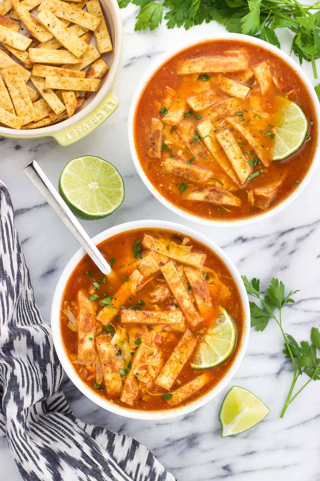 Two bowls of soup topped with tortilla strips, lime wedges, and cheese ready to eat.