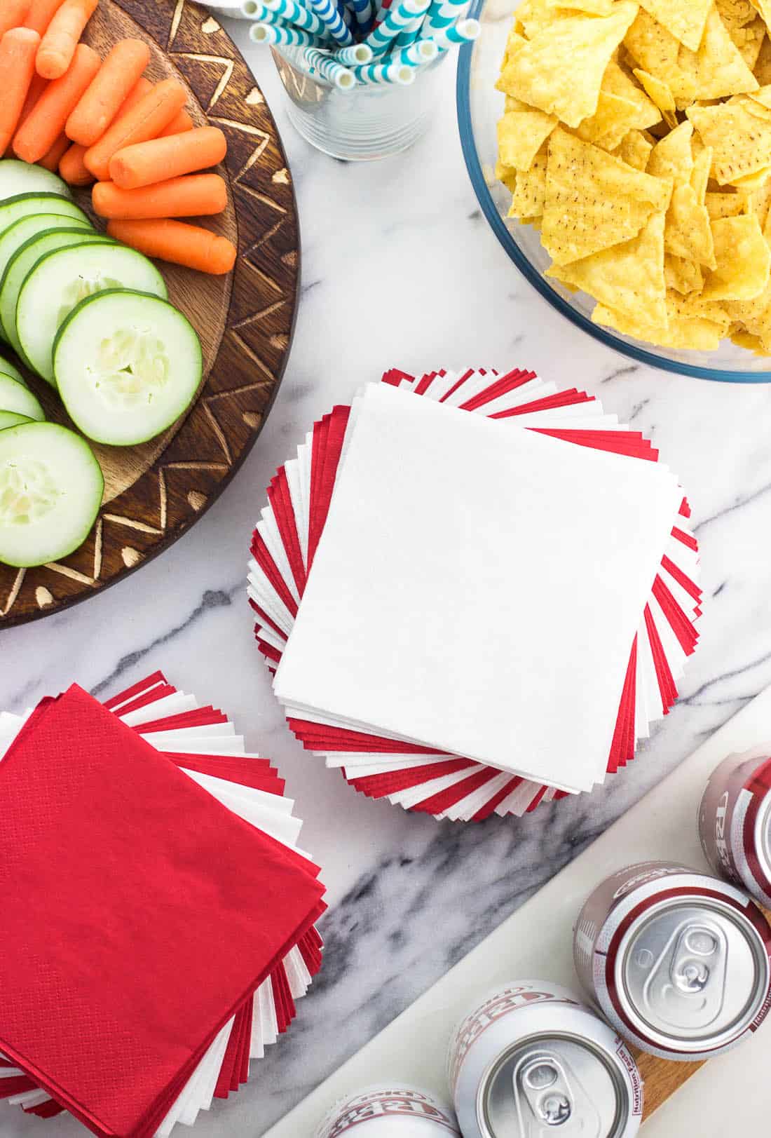 A stack of napkins, a bowl of tortilla chips, and sliced vegetables on a party table.