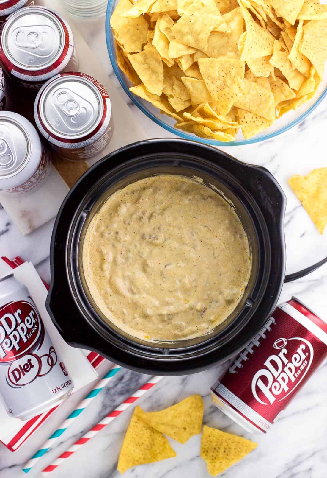 Beef queso dip in a slow cooker next to a bowl of tortilla chips and cans of Dr Pepper.