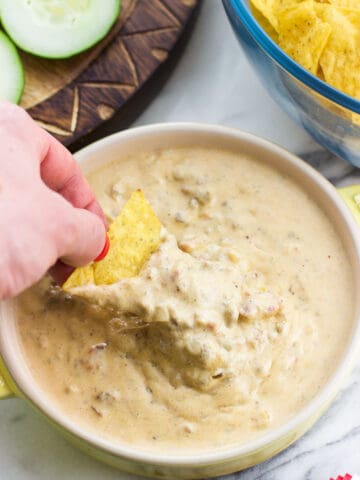 A hand dipping a tortilla chip into a bowl of beef queso dip.