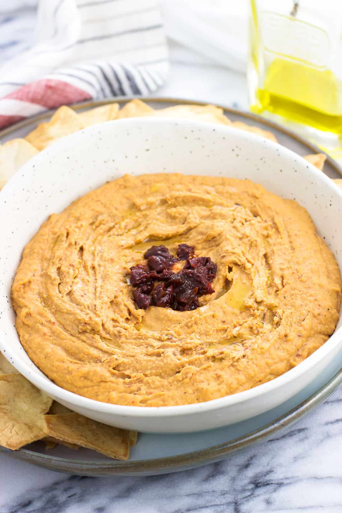 A ceramic bowl filled with hummus, featuring a drizzle of olive oil and chopped chipotle peppers as garnish