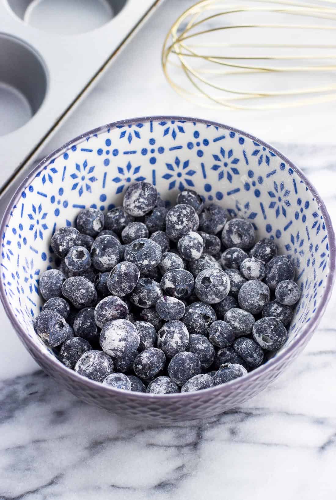 A ceramic cereal bowl filled with flour-dusted blueberries next to a muffin pan and a whisk