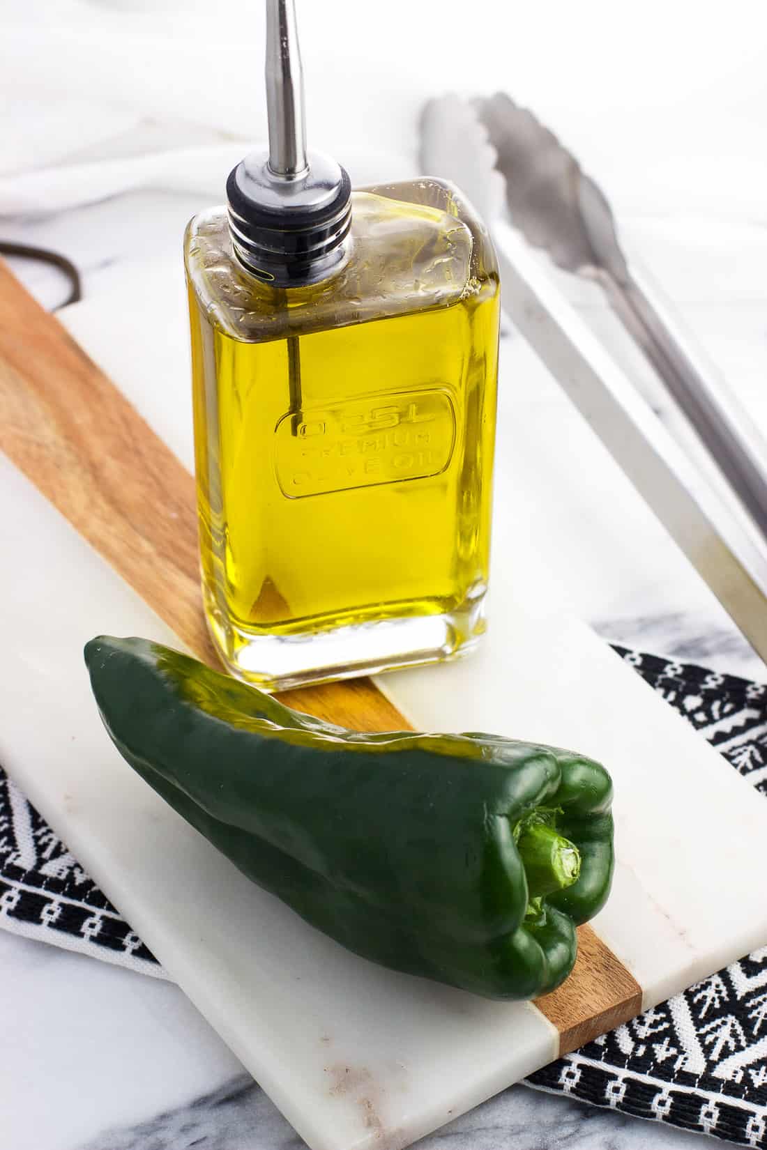 A raw poblano pepper and a glass olive oil dispenser on a marble tray.