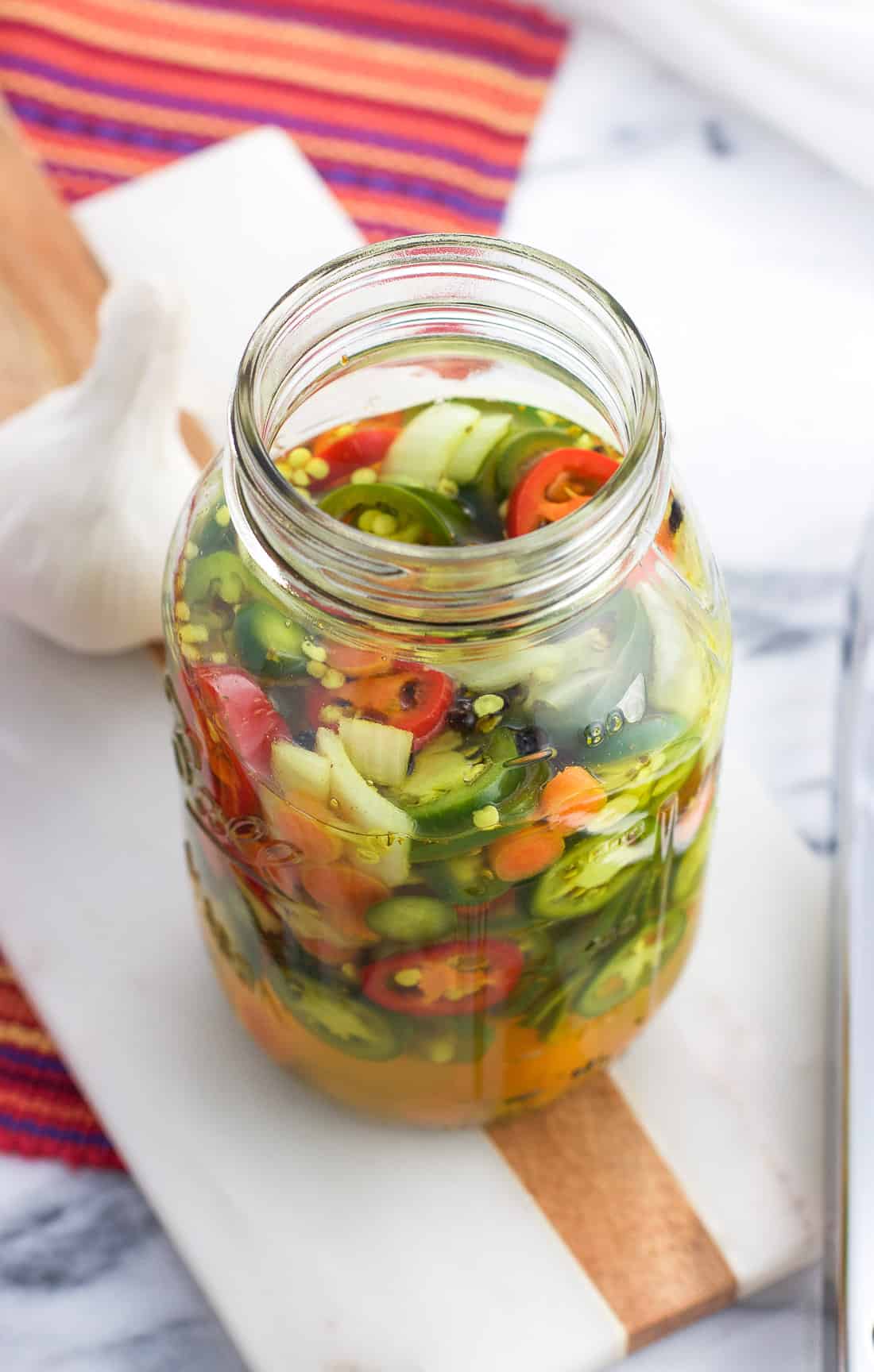 A large glass jar filled with sliced jalapeno peppers, onions, garlic, and carrots and pickling liquid next to a whole head of garlic