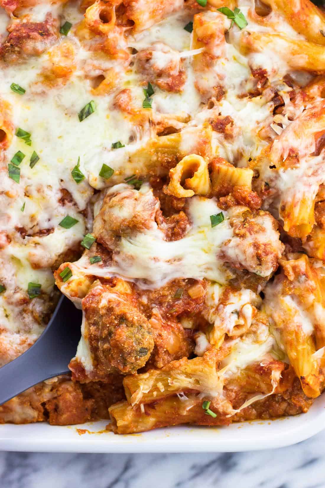 A close-up of a serving spoon lifting out a scoop of baked ziti from the dish.