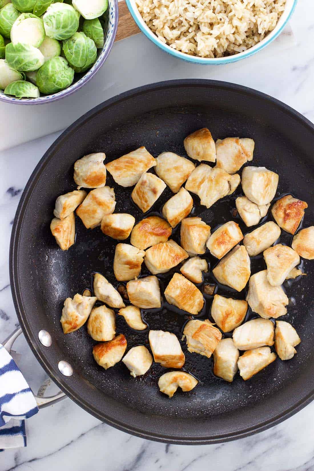 Cooked chicken pieces in a skillet next to bowls of Brussels sprouts and cooked brown rice.