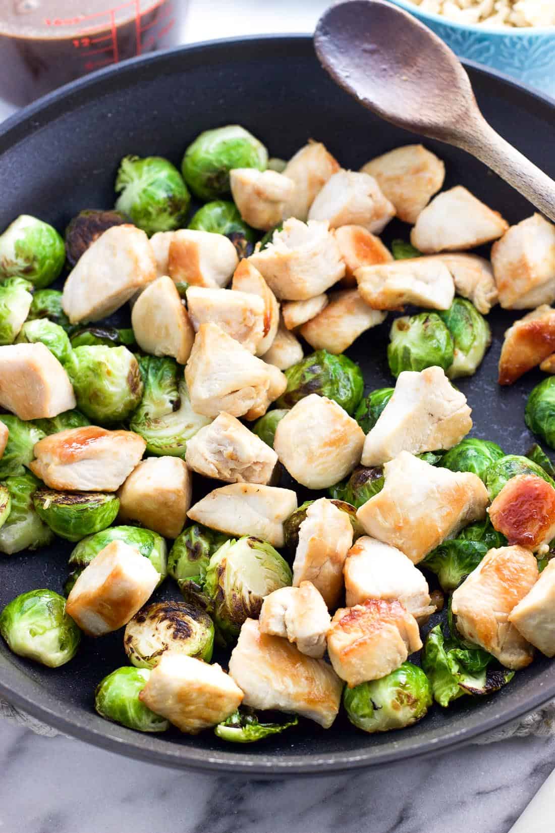 Cooked chicken and sauteed Brussels sprouts in a skillet next to a wooden spoon.