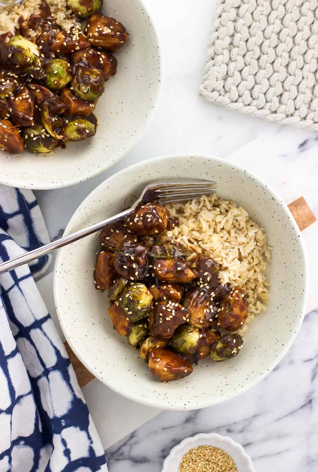 Two bowls of stir fry garnished with sesame seeds served alongside brown rice