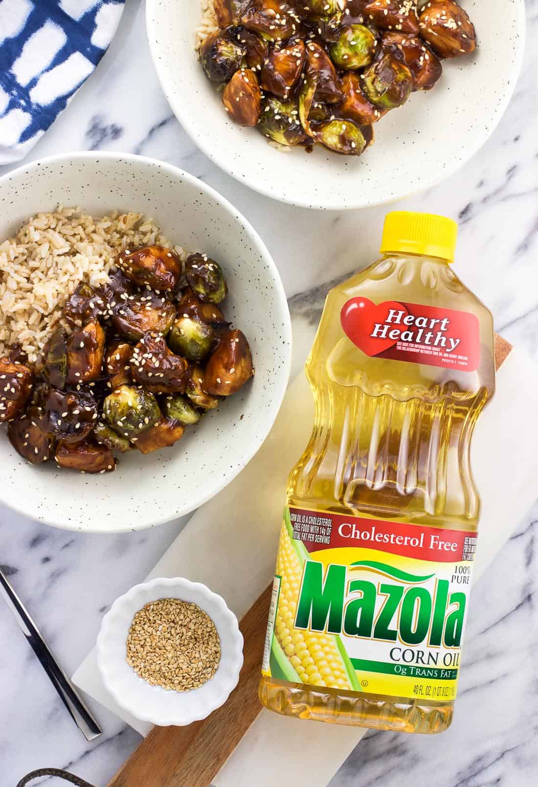 A bottle of Mazola corn oil next to two plates of chicken stir fry served with brown rice and a small bowl fo toasted sesame seeds