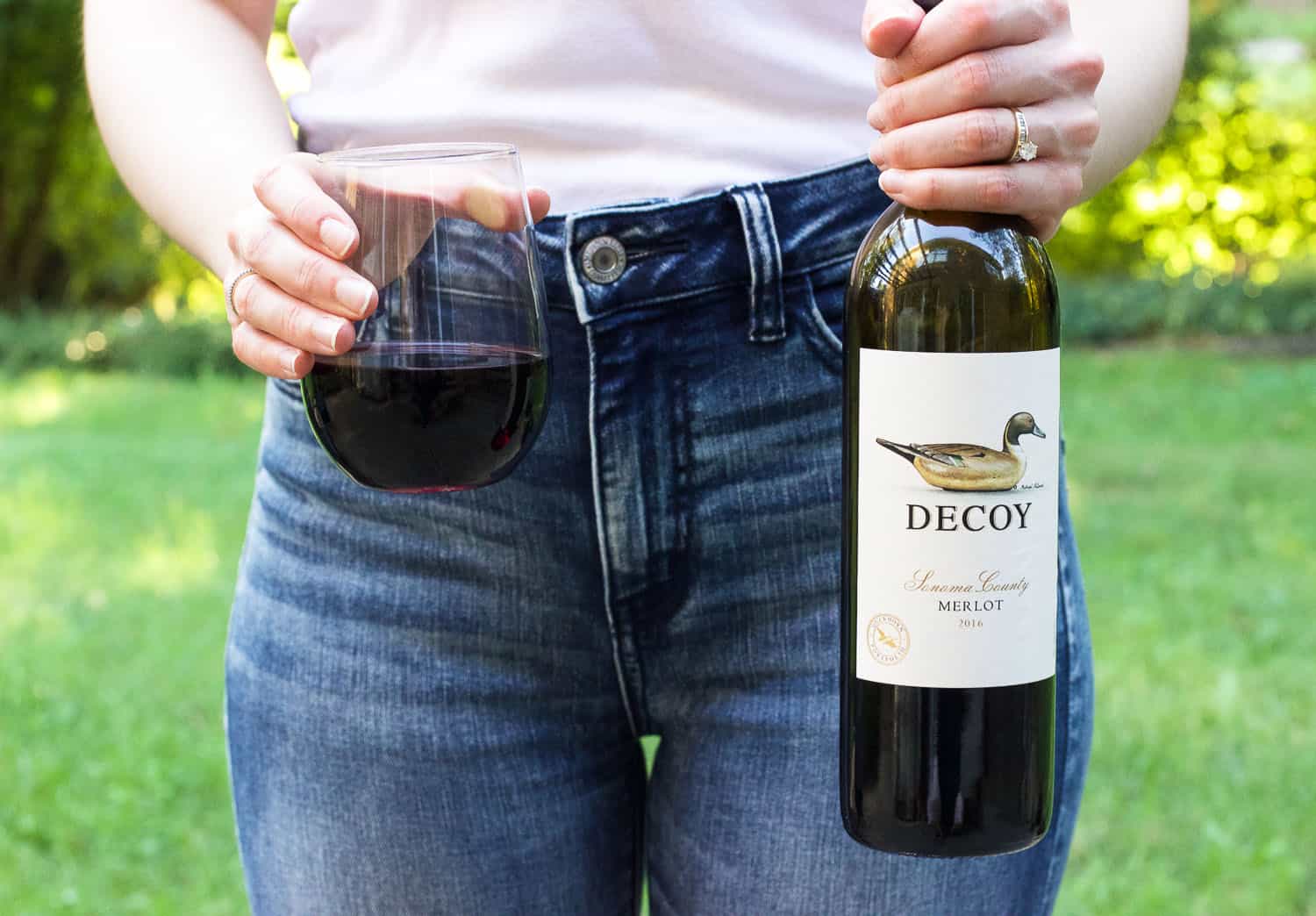 A woman standing outside holding a bottle of Decoy Merlot in her left hand and a stemless wine glass of the wine in her right