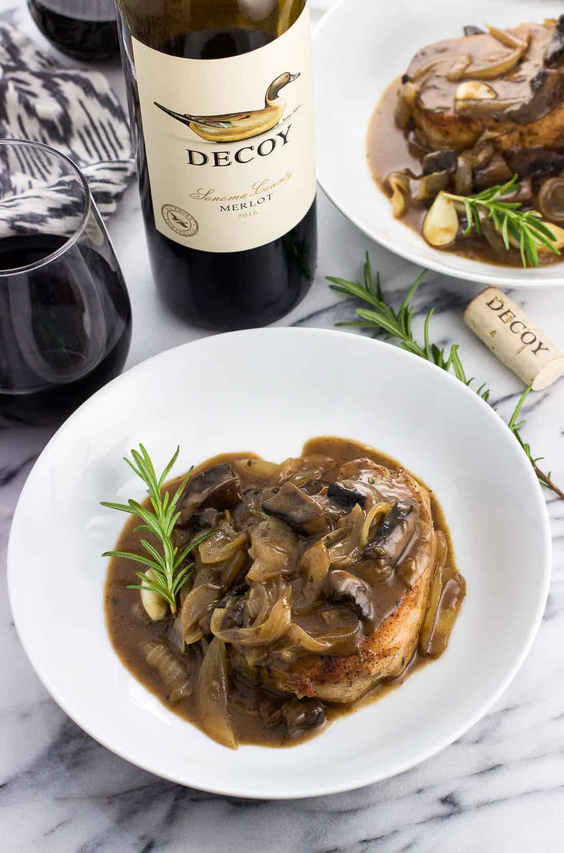 A pork chop covered in sauce on a plate with a rosemary sprig next to a glass and bottle of wine.