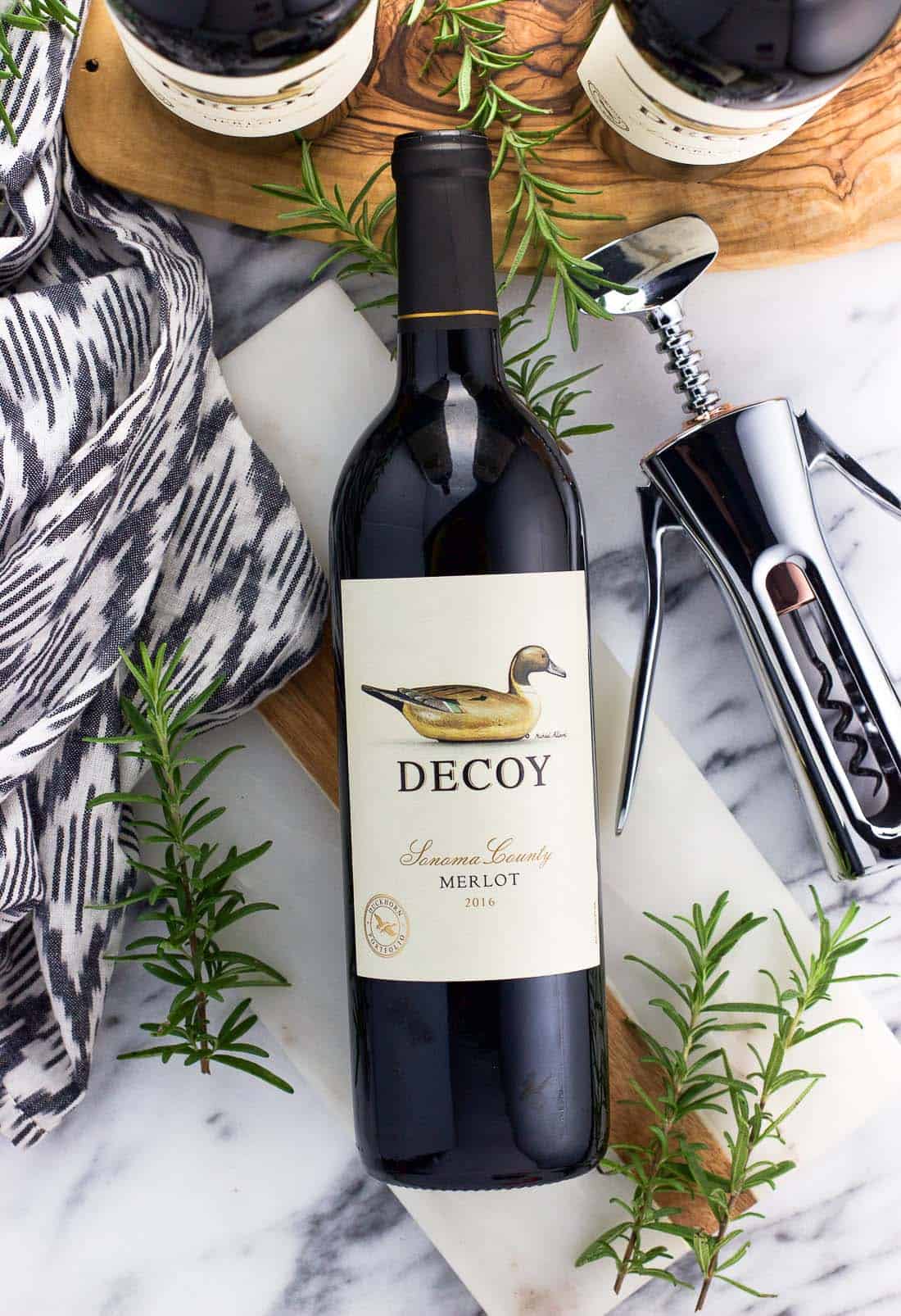 An overhead shot of a bottle of Decoy Merlot, surrounded by two more bottles, a metal wine opener, and rosemary sprigs