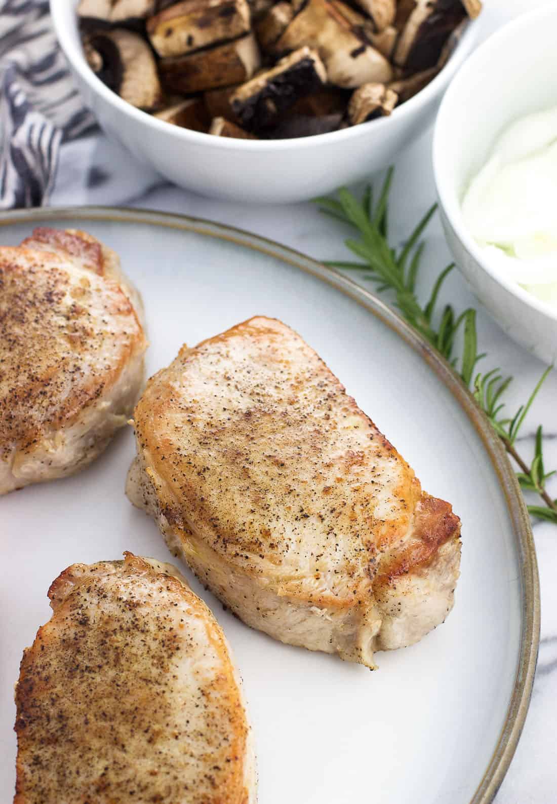 Three seared pork chops on a plate surrounded by bowls of mushrooms and sliced onions.