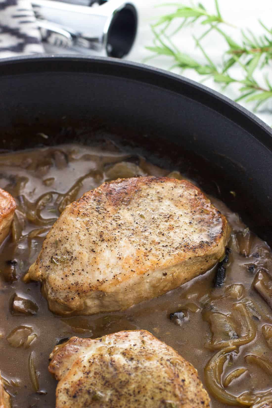 Seared pork chops in a skillet with sauce, mushrooms, and onions.