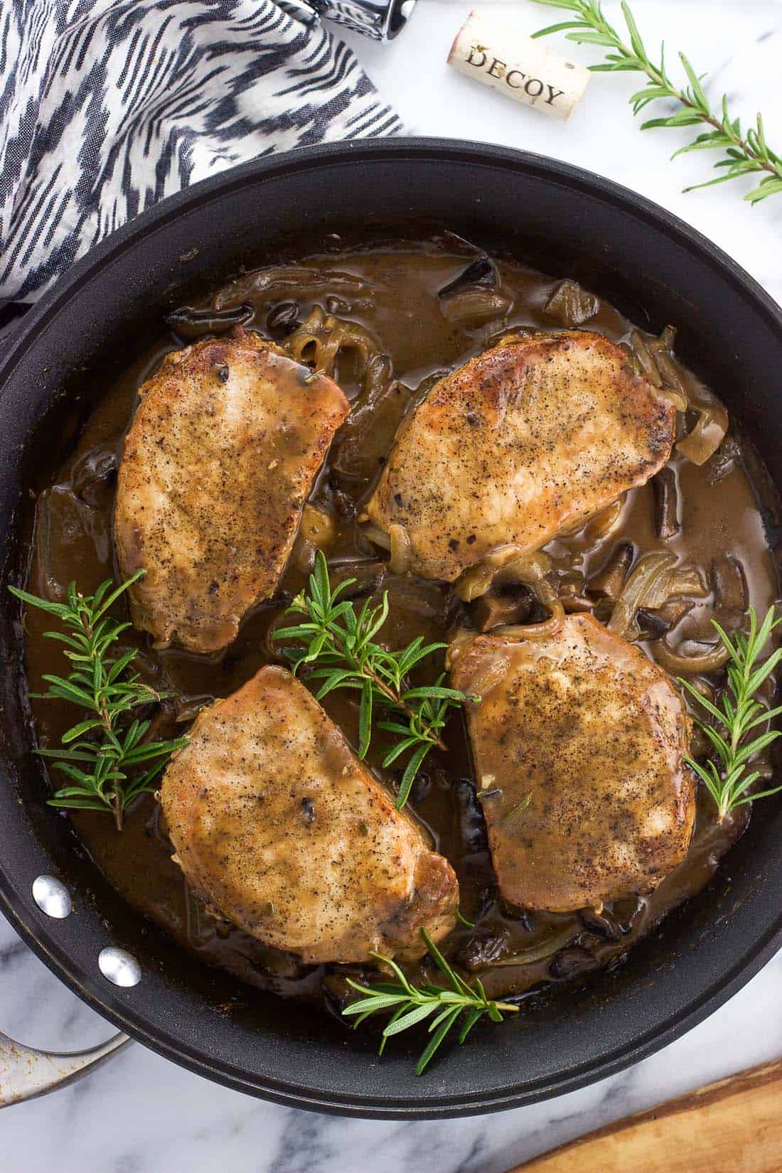 An overhead shot of four cooked pork chops in a skillet with sauce and rosemary sprigs, all ready to serve.