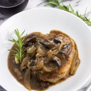 A pork chop covered in sauce on a plate with a rosemary sprig next to a glass of wine.