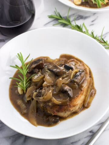 A pork chop covered in sauce on a plate with a rosemary sprig next to a glass of wine.