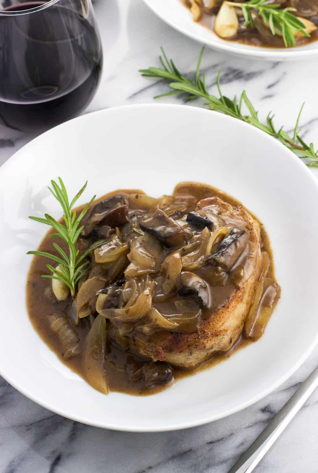 A pork chop covered in sauce on a plate with a rosemary sprig next to a glass of wine.