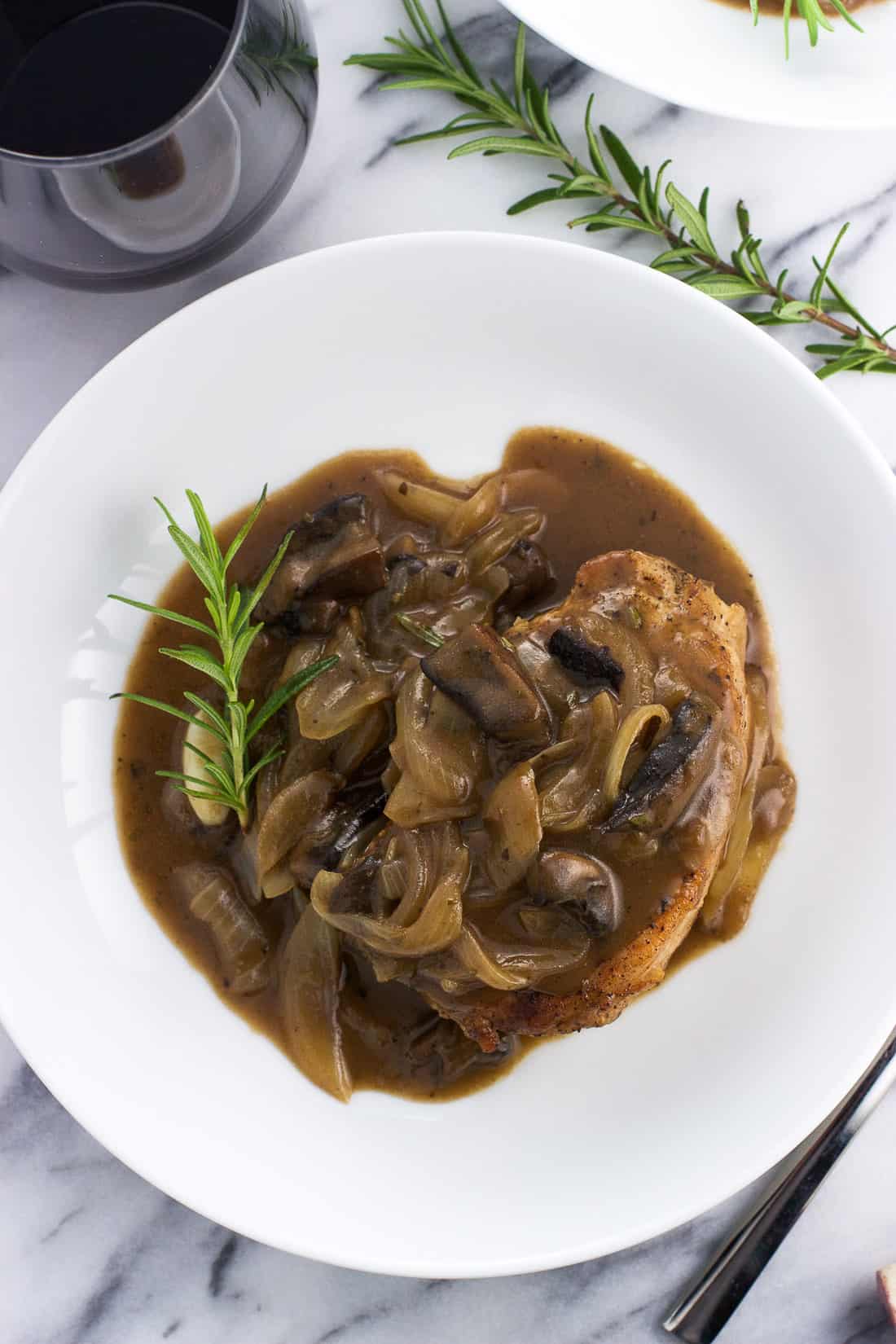 An overhead picture of a serving of pork chops and sauce in a shallow dinner plate next to a glass of Merlot.