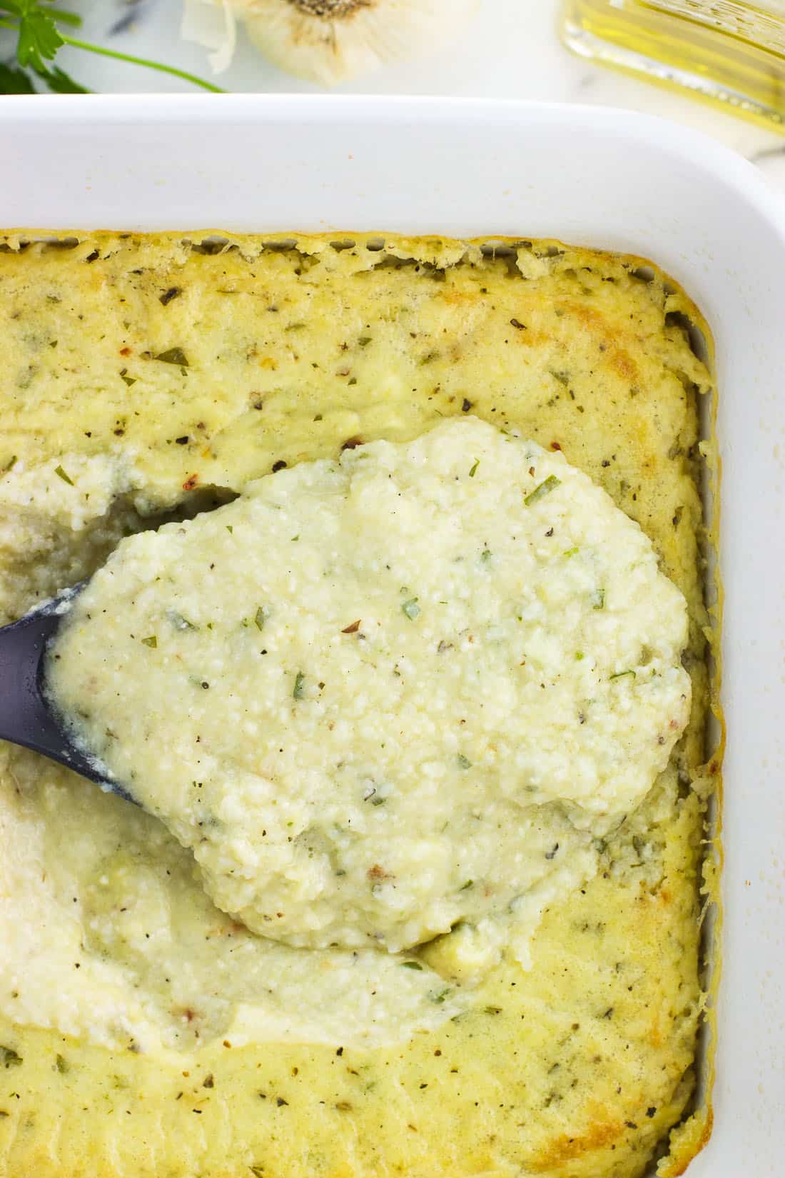 A serving spoon lifting out a scoop of the baked grits from the baking dish