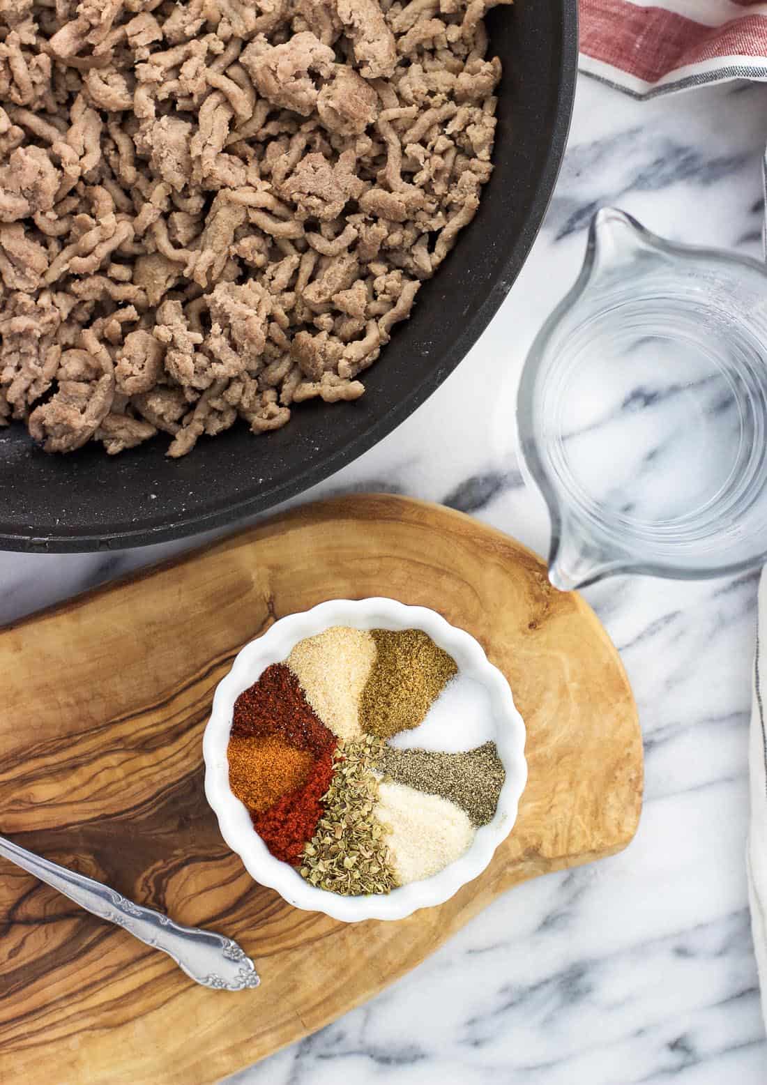 A skillet of cooked ground beef next to a cup of water and a bowl of taco seasoning.