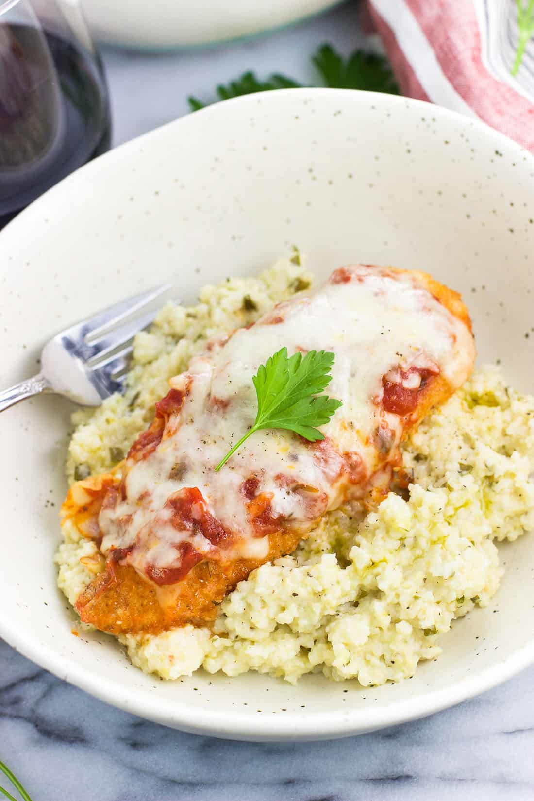 A breaded chicken cutlet covered in marinara sauce and cheese served over grits in a shallow bowl with a fork